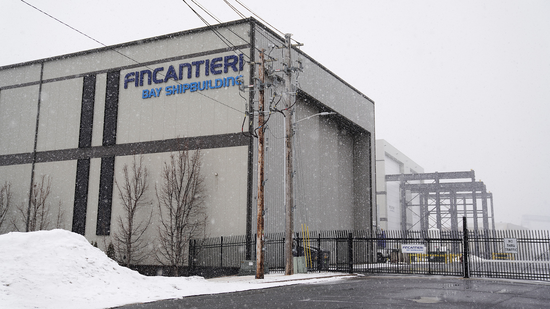 Snow flies around two multi-story buildings, connected to power lines and cranes, with one in the foreground bearing the wordmark "Fincantieri Bay Shipbuilding."