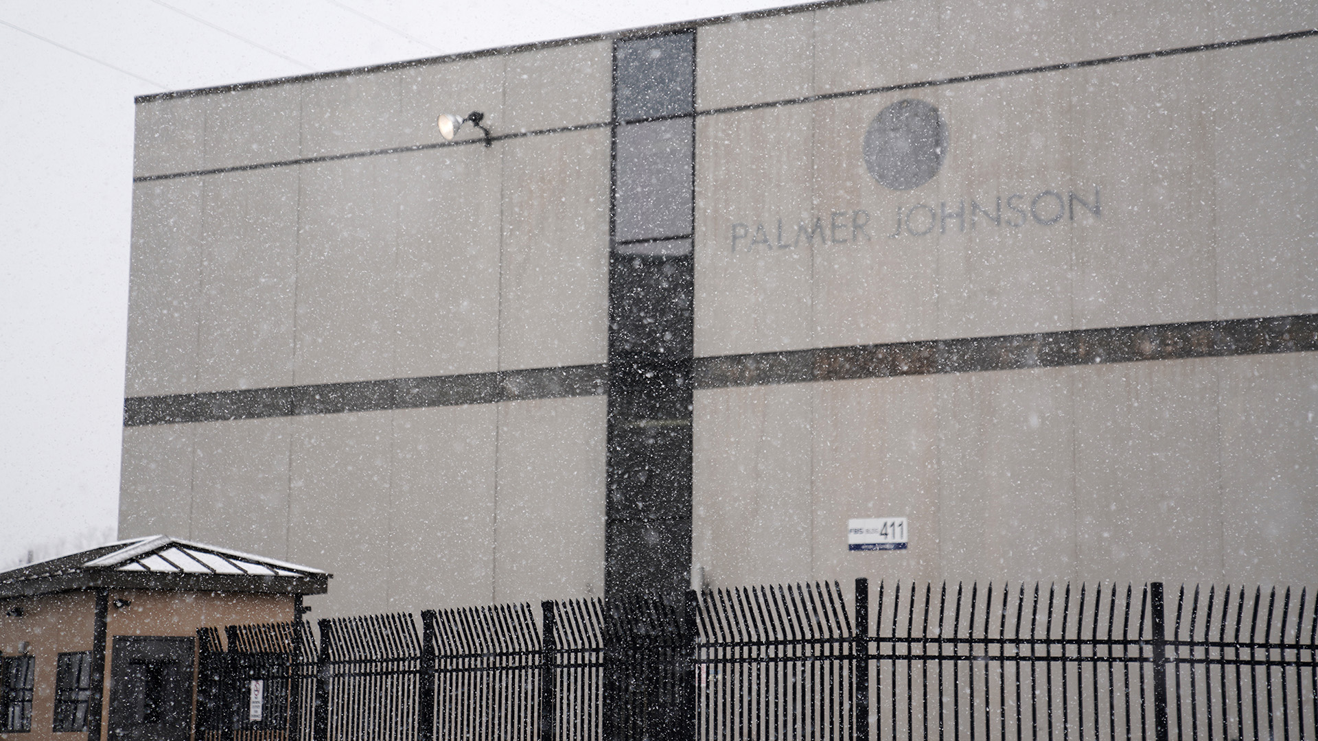 A multistory building is marked by discoloration where a logo and wordmark stating "Palmer Johnson" were formerly placed, with a guardhouse and wrought iron fence in the foreground and snow flying in the air.