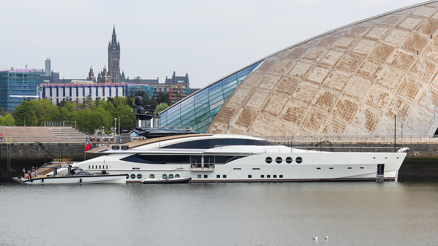 A yacht in calm waters is docked next to a modernist building with a city skyline in the background.