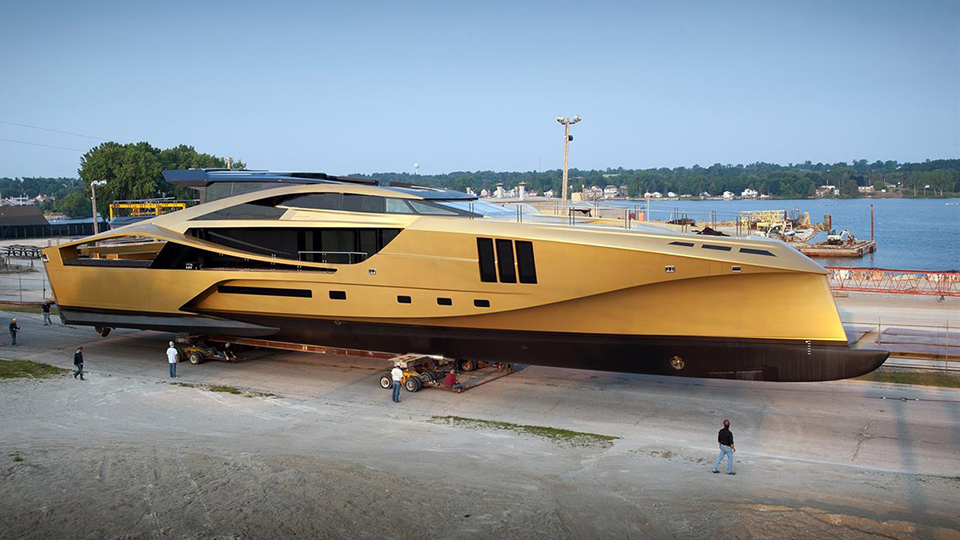 A yellow luxury yacht sits on a trailer on a road next to a waterway, with the opposite shore in the background.