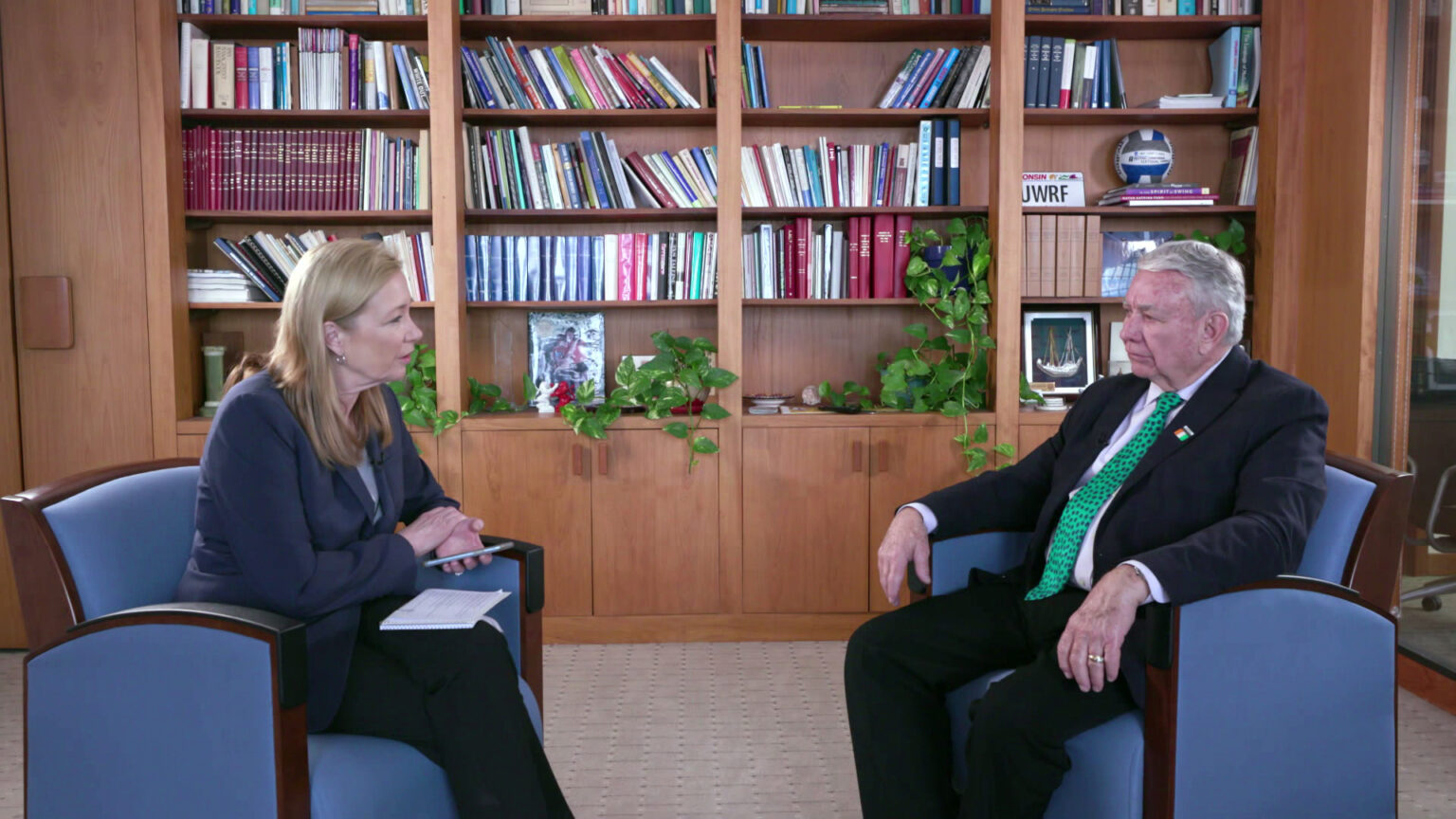 Frederica Freyberg and Tommy Thompson sit in chairs in a room with a bookshelf in the background.