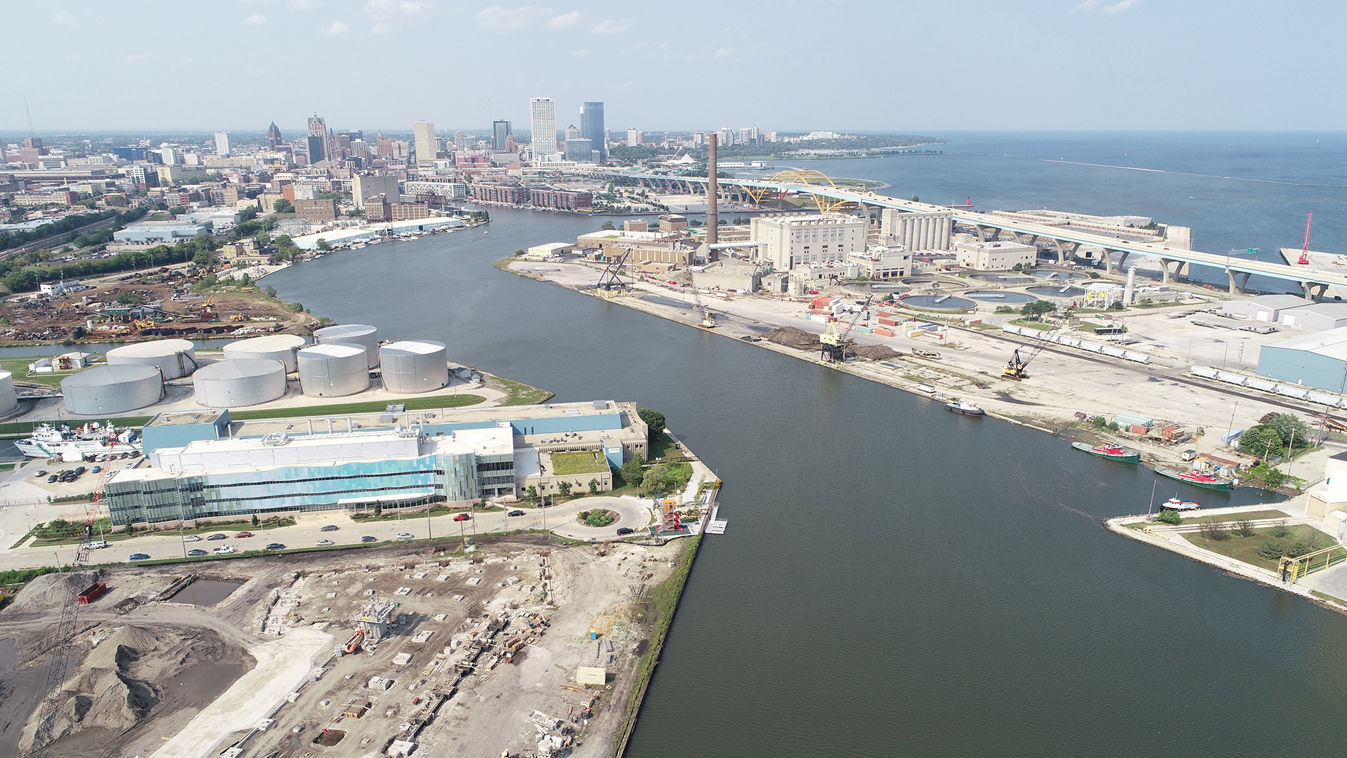 An aerial photo shows a wide river mouth surrounded by industrial development, with a downtown skyline in the background.