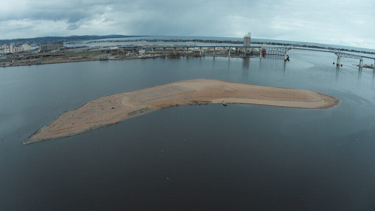 A island with a dirt surface is surrounded by calm waters, with a bridge, cityscape and more water in the background.