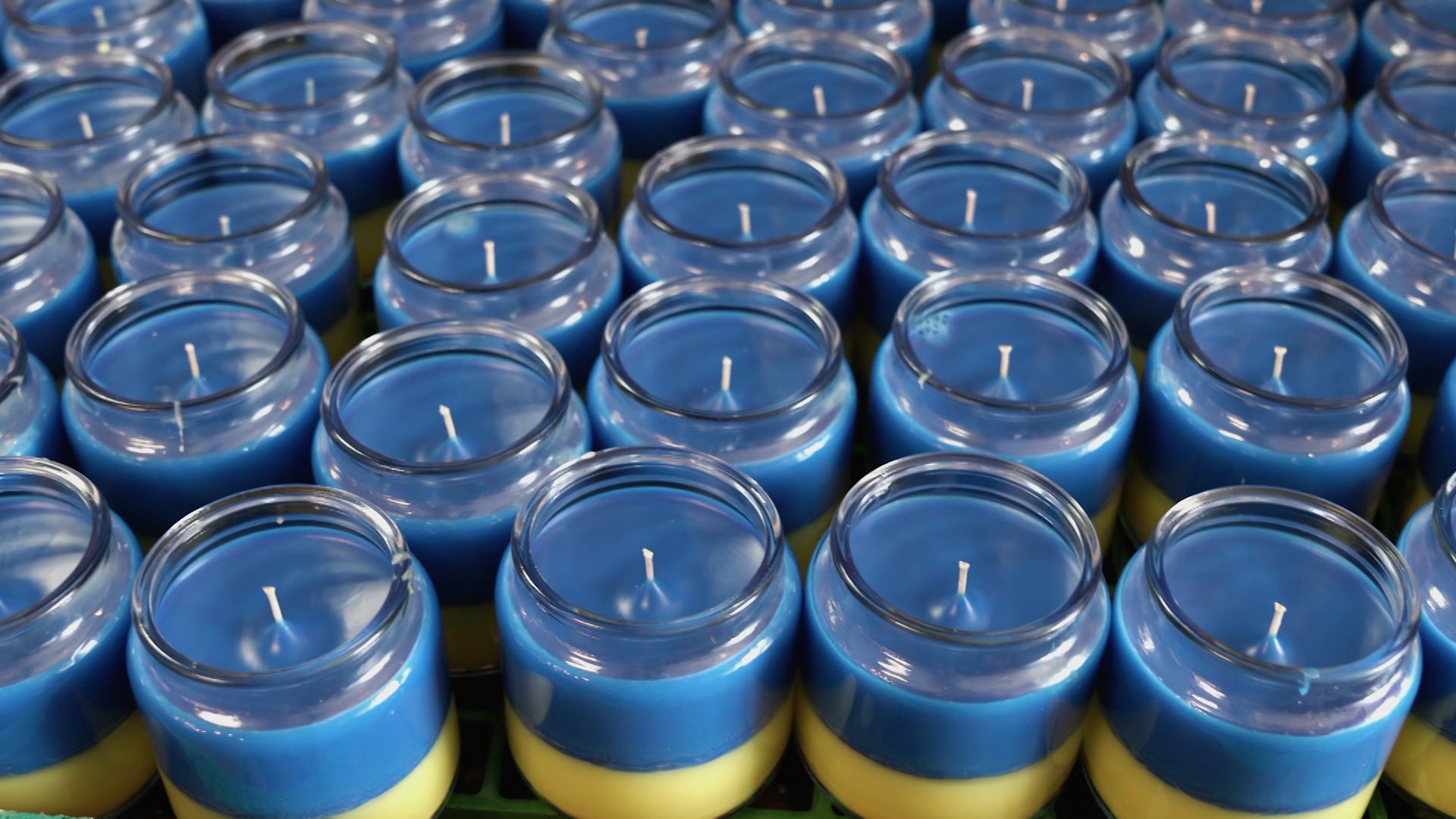 Rows of candles in glass jars with a top blue layer and a bottom yellow layer sit on a table.