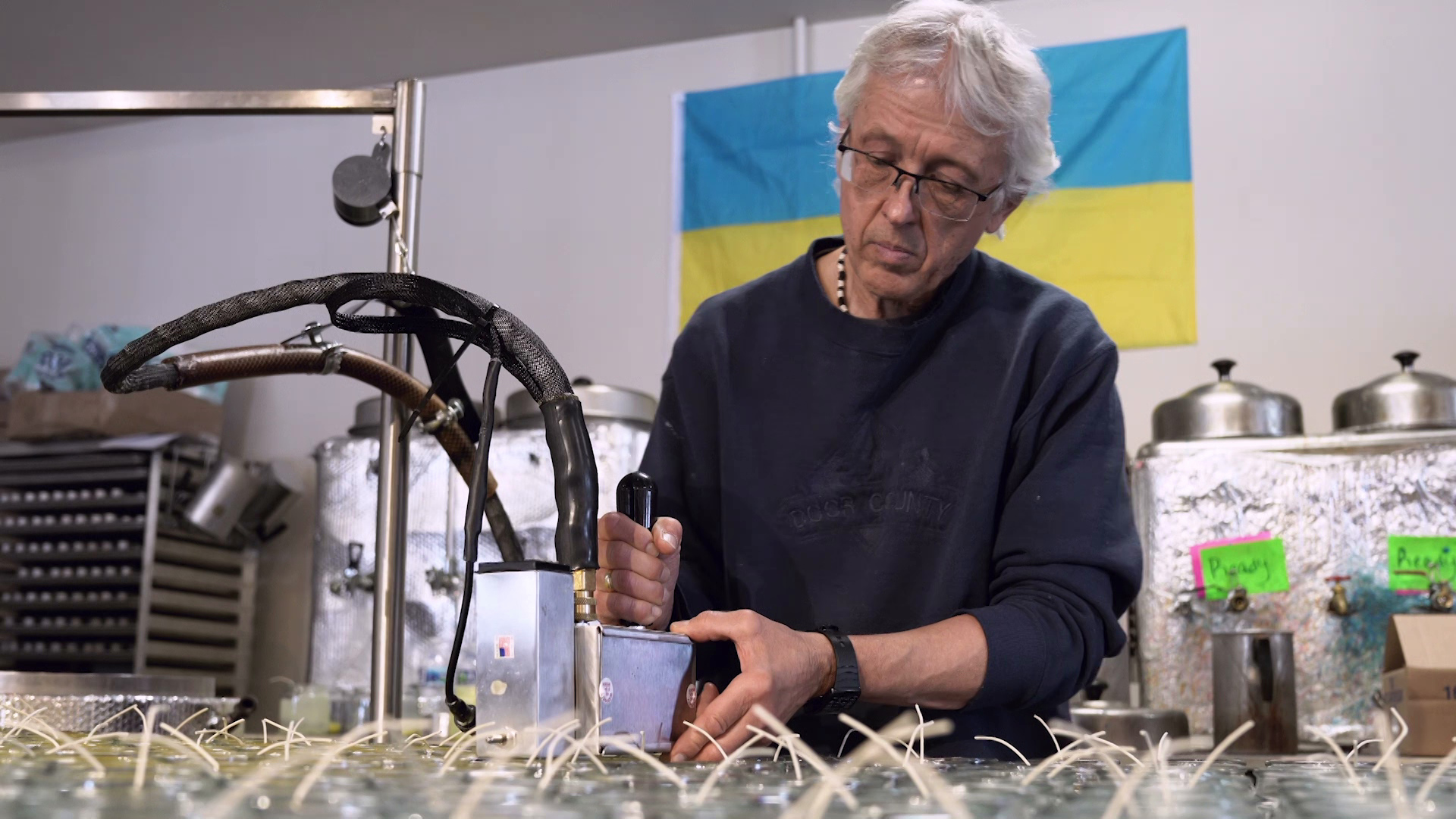 Dr. George Gorchynsky uses a machine to work with wax, with rows of empty candle jars with wicks in the foreground and a Ukrainian flag pinned to the wall in the background.