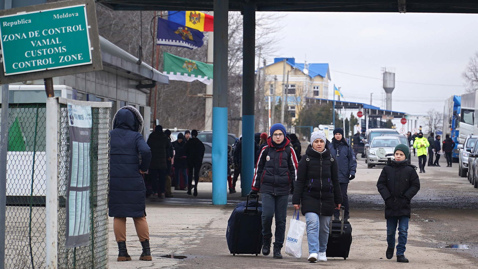 A trio of people, two who carry bags and rolling luggage, walk through a border customs control zone, with other people and vehicles lined up in the background.
