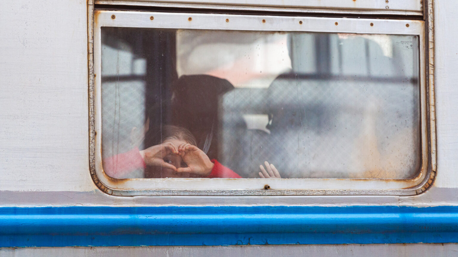 A child inside a train looks out a window and makes a heard symbol with their hands.