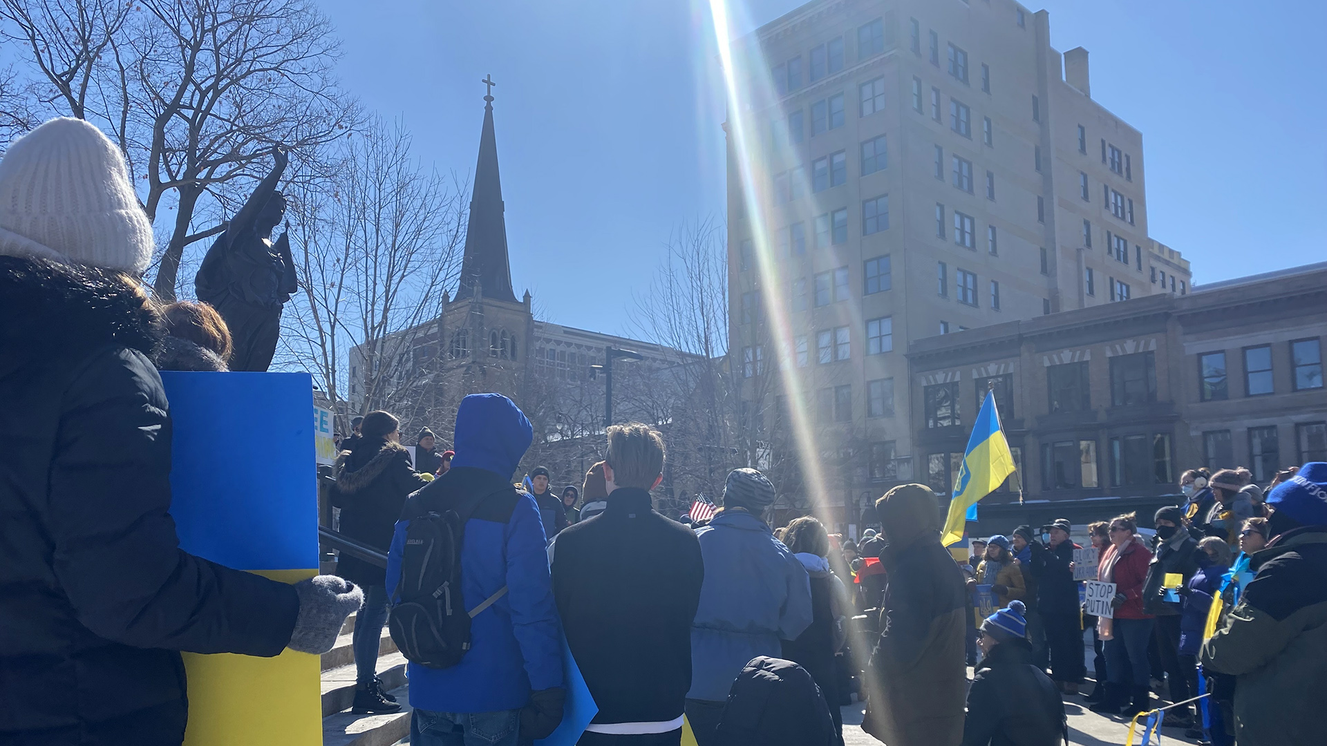 A sunbeam lens flare shines over an outdoor rally next to a statue, with protestors holding blue-and-yellow signs and flags, with buildings in the background.