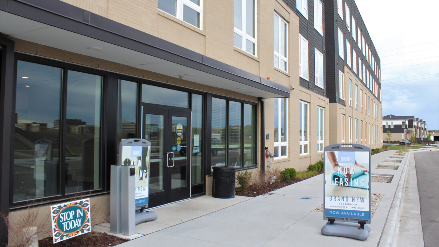 Sidewalk signs that read Now Leasing stand in front of the door to a newly constructed, four-story apartment building.