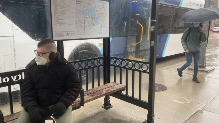 A masked passenger disembarks from a bus while another passenger wearing a mask sits at a bus stop