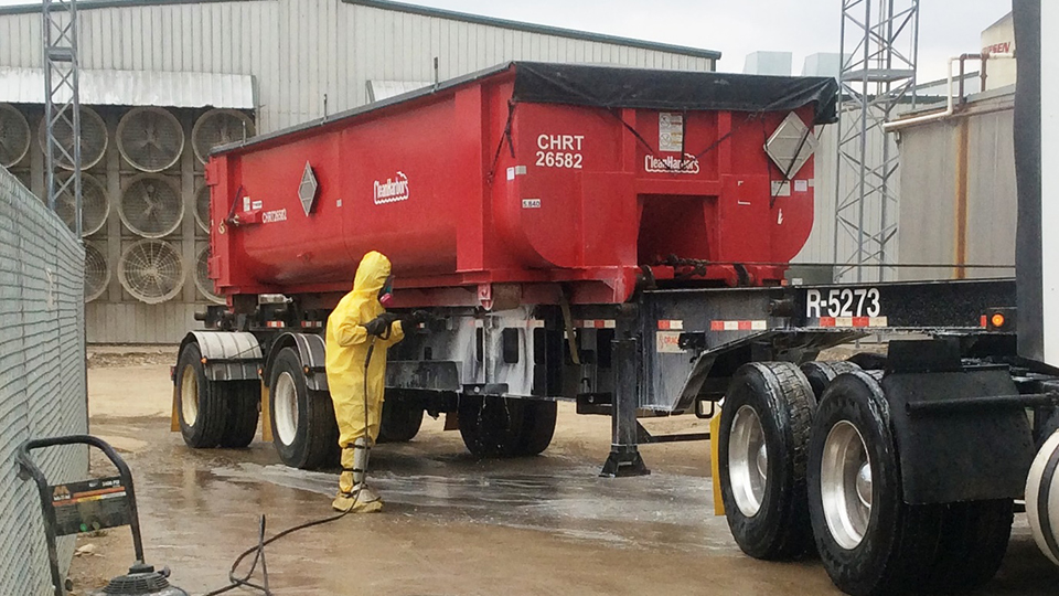 A person wearing a hazmat suit uses a high-pressure hose to wash the undercarriage of a truck trailer topped with an open bed.