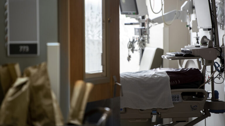 A hospital bed surrounded by medical equipment sits empty as seen through a door.