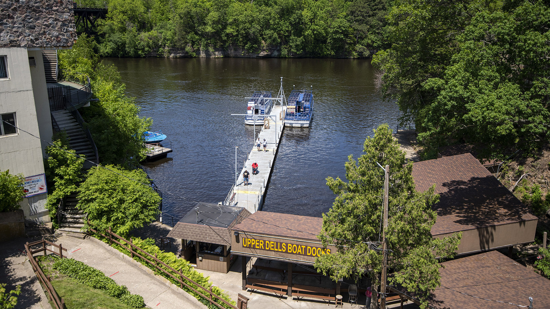 Excursion boats are docked on a pier extending from a building with the sign "Upper Dells Boat Docks," with other buildings on the near side of the river and trees covering the opposite shores.