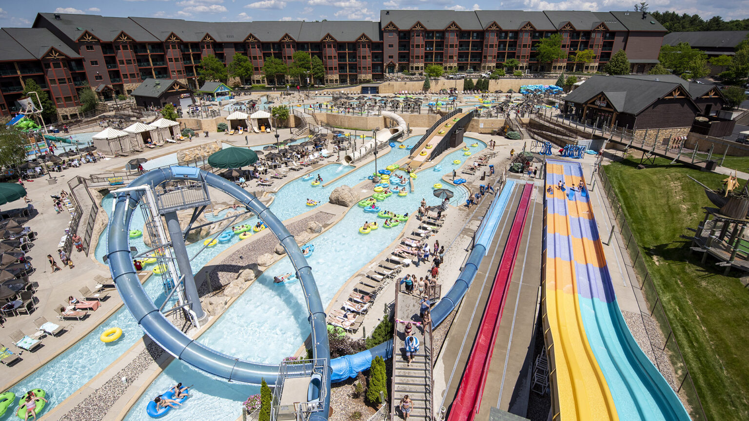 Tourists go down waterslides and float on tubes in a lazy river in an outdoor waterpark, with multistory hotel buildings in the background.