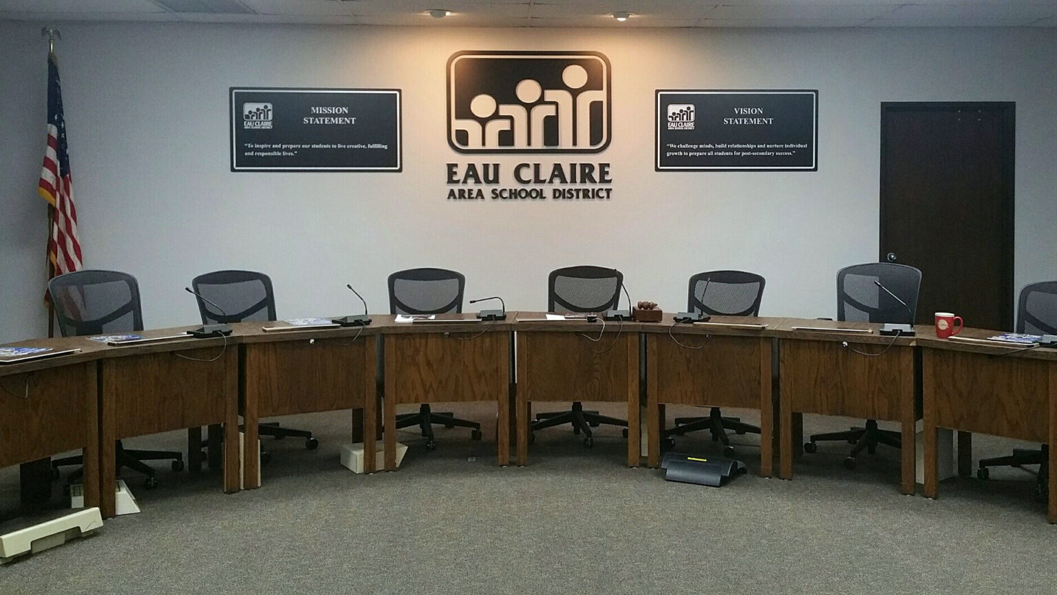 A meeting room with an arc of wood desks is backed by empty chairs and a sign on the wall reading Eau Claire Area School District along with a mission statement and a vision statement.