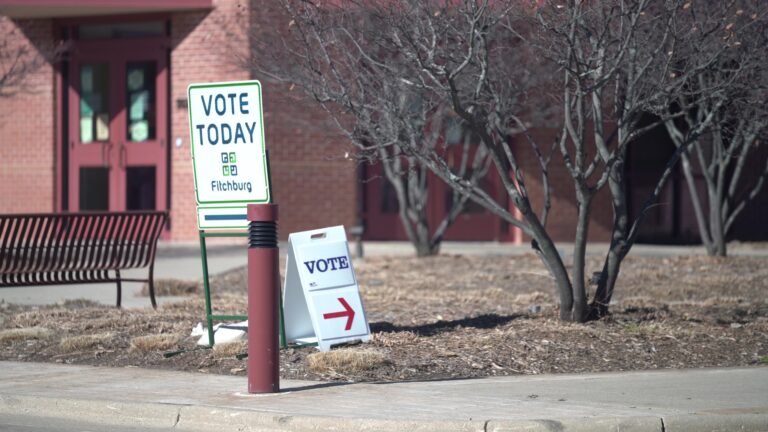 Two signs stating Vote Today and Vote with arrows stand amid leafless trees and a bench in front of a brick building.