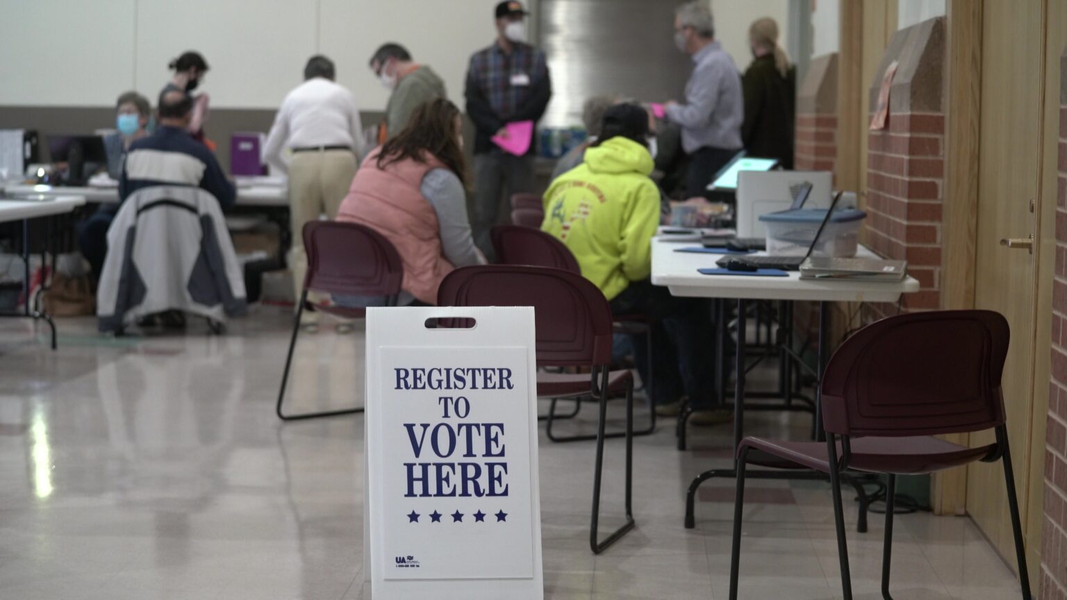 A sign stating Register to Vote Here stands in a large room with people standing and seated at tables in the background.