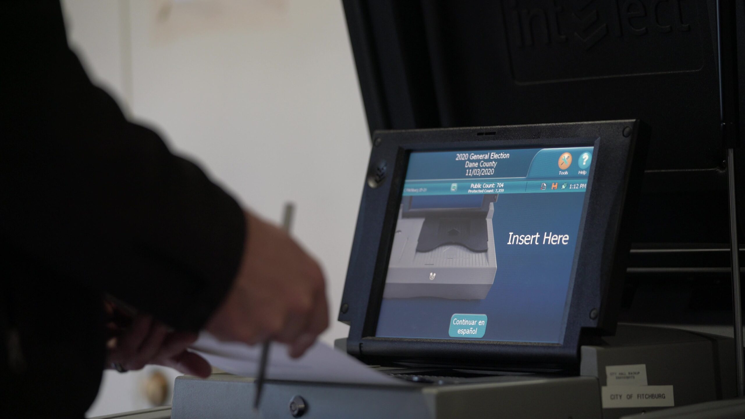 A voting tabulation machine with a touch screen graphic shows instructions while a voter enters a paper ballot into it.