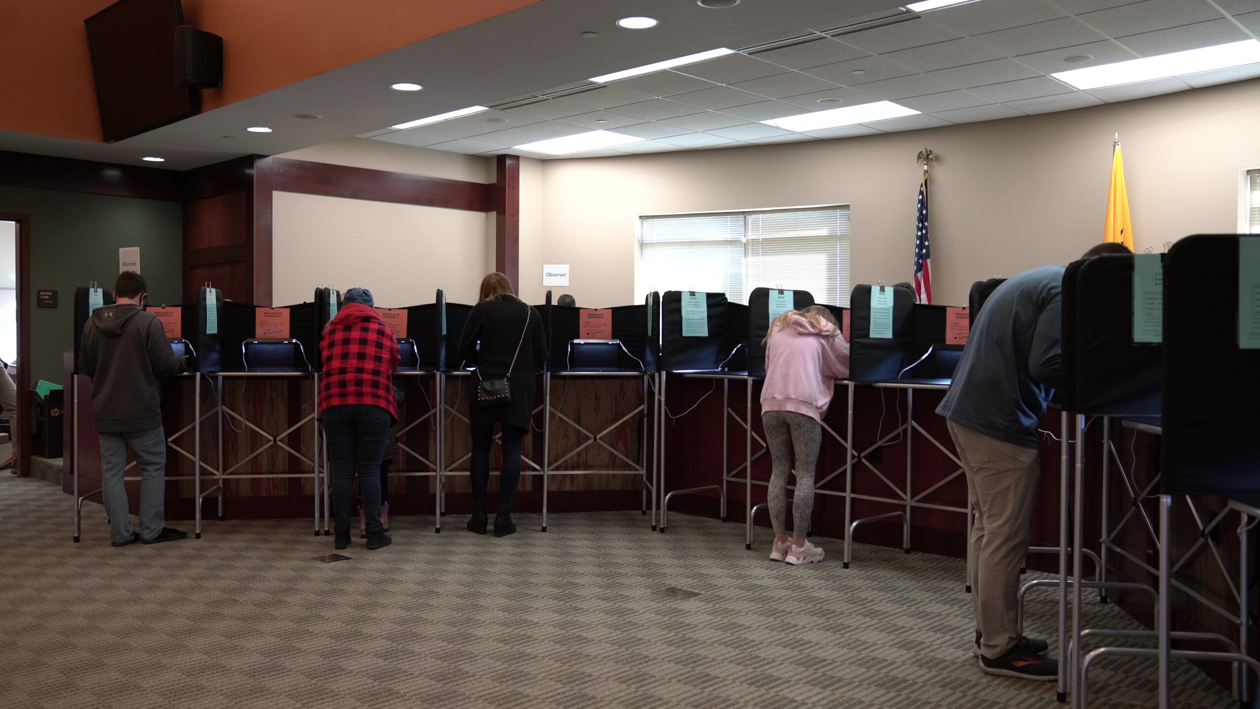 People stand and vote in booths with metal legs and vertical privacy dividers in a government building with a U.S. flag.