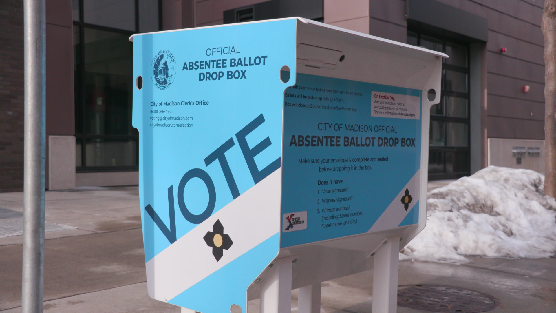 A metal box with a thin slot and labels stating "VOTE" and "City of Madison Official Absentee Ballot Drop Box" stands on a sidewalk, with a pile of snow and buildings in the background.