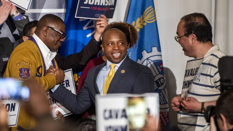 Cavalier Johnson grasps the hand of one supporter and smiles at another while standing in a crowd in front of the flags of Wisconsin and Milwaukee.