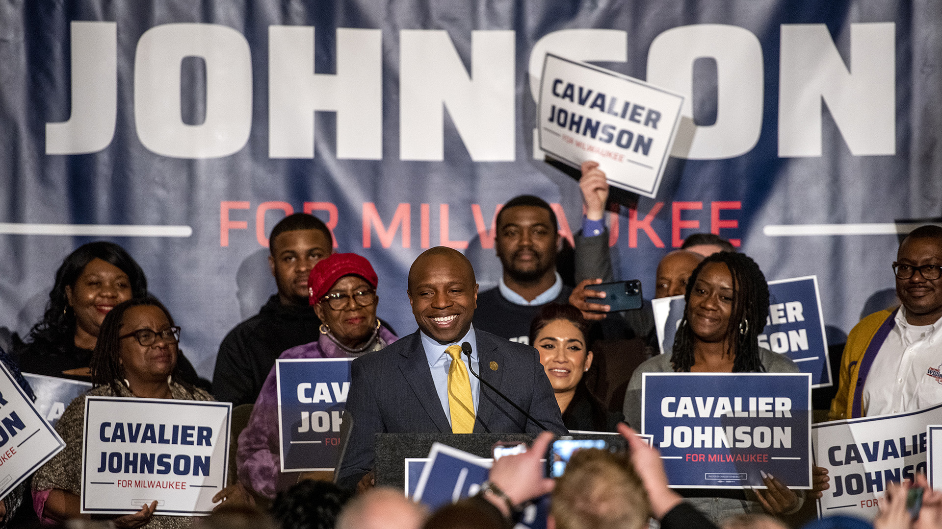 Cavalier Johnson stands at a podium in front of a group of supporters holding "Cavalier Johnson for Milwaukee" campaign signs.