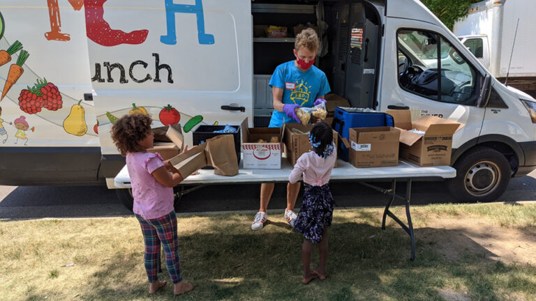 Two children stand in front of a table stacked with food boxes set up next to a large van parked on the side of a road.