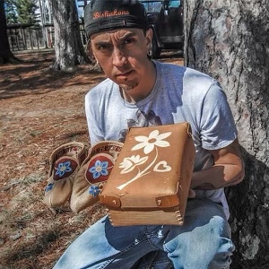 Greg Biskakone Johnson holds moccasins and a box while seat on a tree.