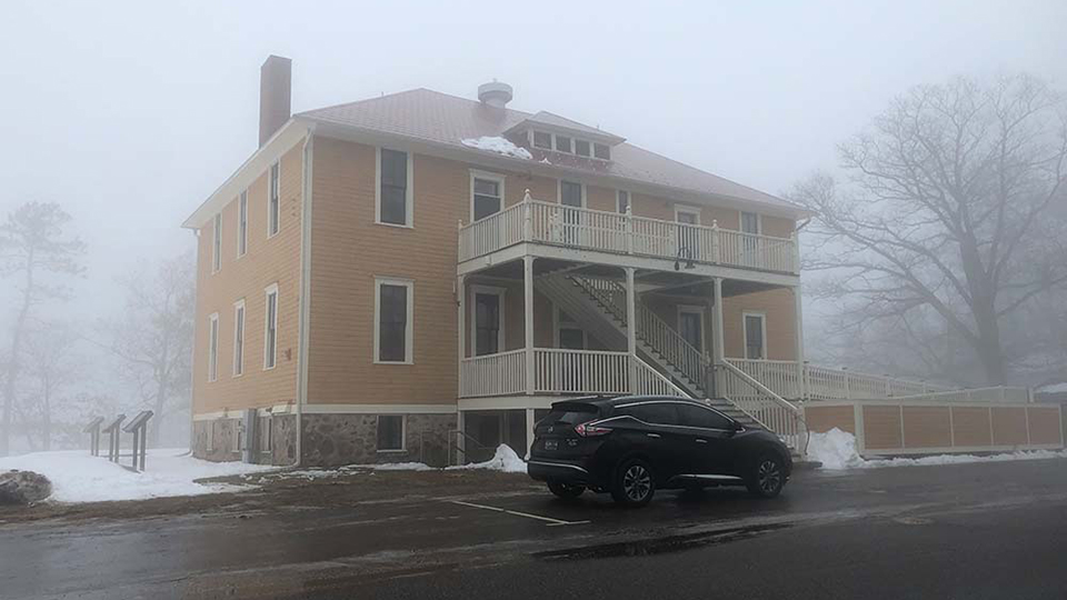 An SUV is parked in front of a three-story building with a stone foundation and wood siding, with snow on the ground and surrounding fog.