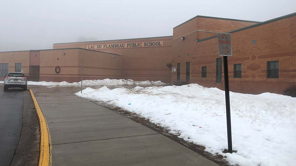 A car is parked next to a brick building with the sign "Lac Du Flambeau Public School," with snow on the ground and fog in the air.