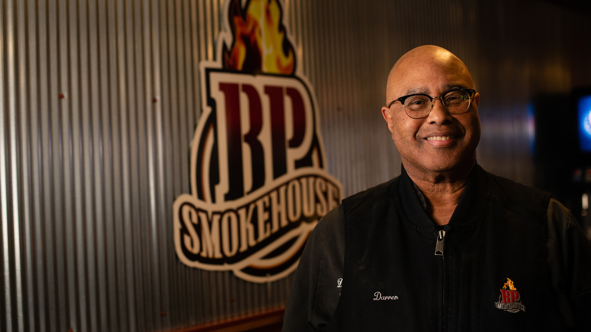 Darren Price smiles for a photo while standing in front of metal wall panels with a BP Smokehouse logo.