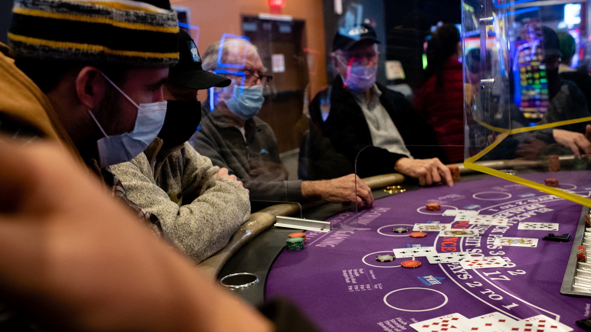 Several people wearing face masks sit at a blackjack table with cards and chips.