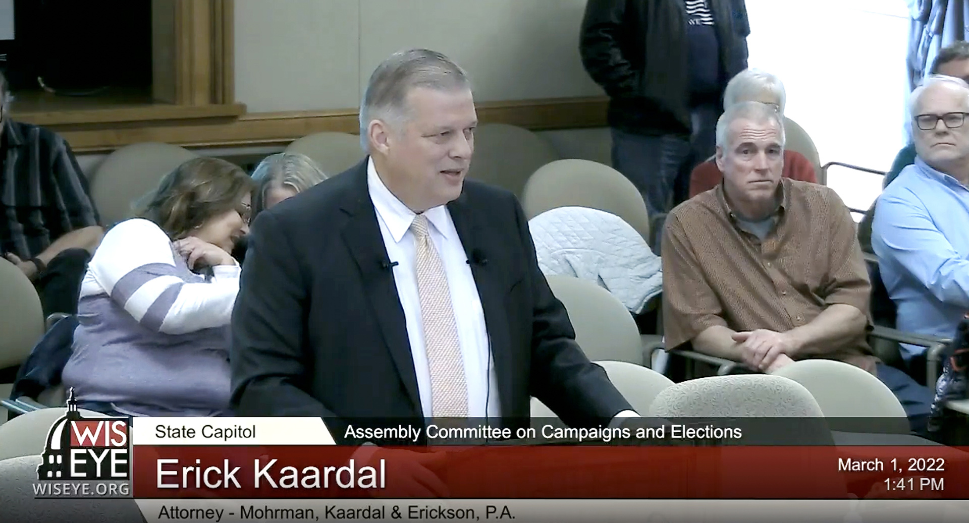A screenshot shows Erick Kaardal speaking in front of a podium in a legislative hearing room, with observers seated in the background.