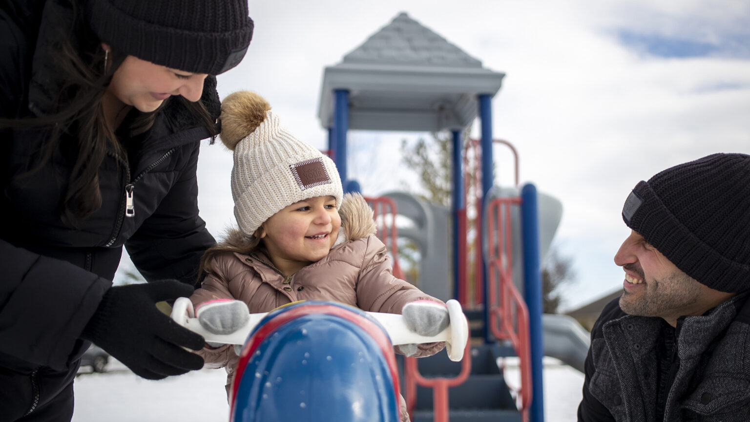 Jennifer Gonzalez and Mario Gonzalez stand and crouch, respectively, on either side of their daughter, who is seated on playground equipment with snow on the ground.