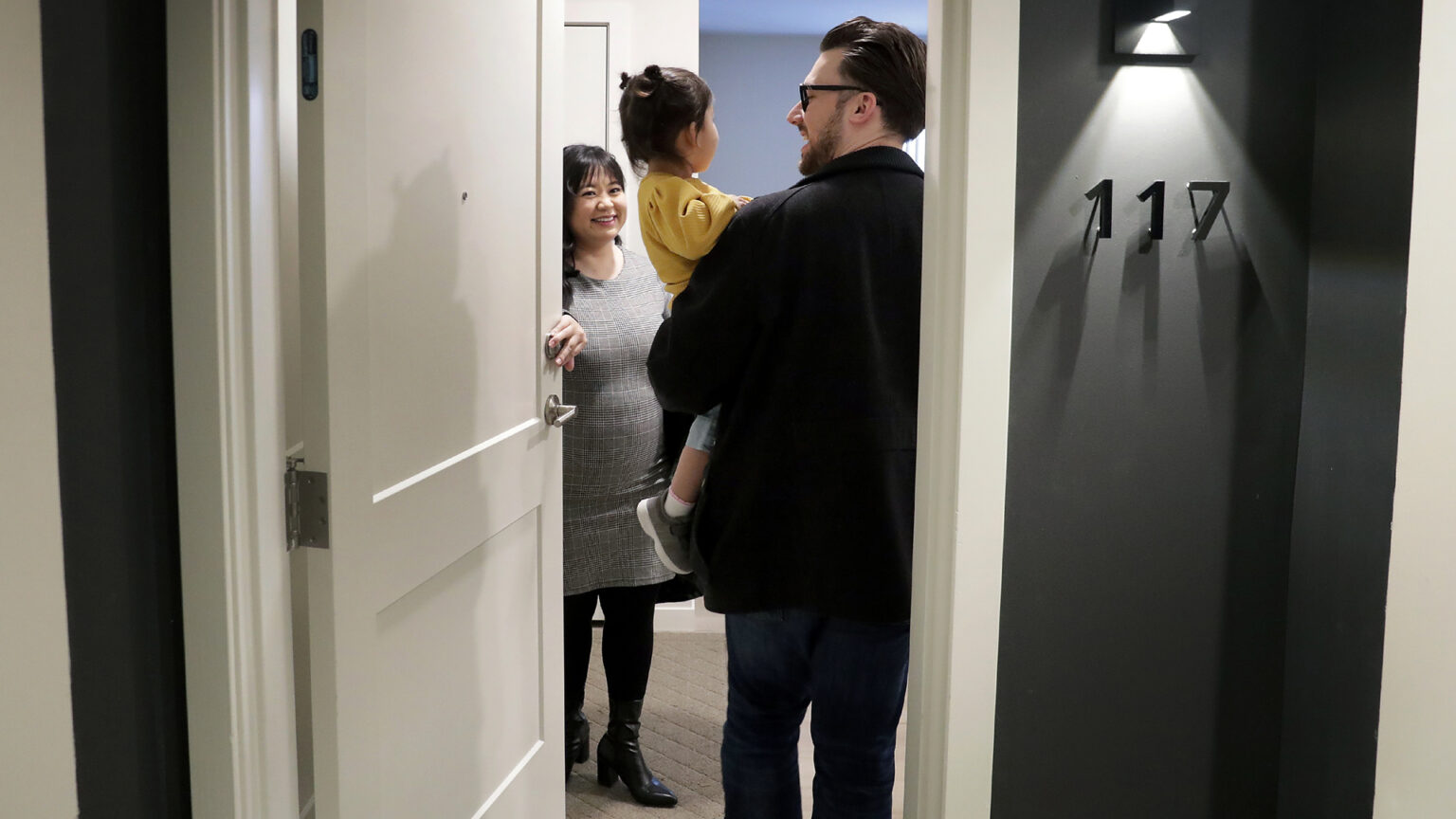 Sheng Lee Riechers stands inside a door to an apartment, facing Karl Riechers, who is holding their daughter.