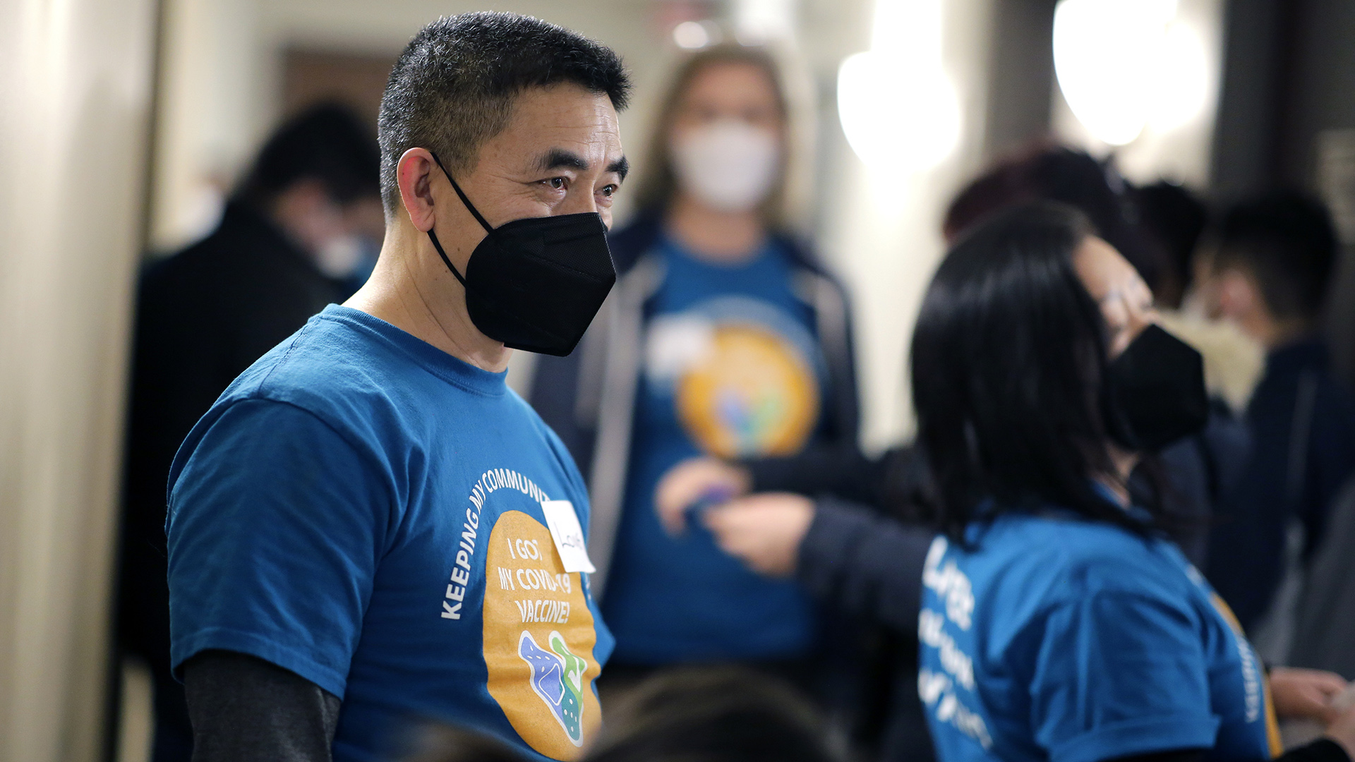 Long Vue stands in a room while wearing a face mask, surrounded by other people wearing face masks.