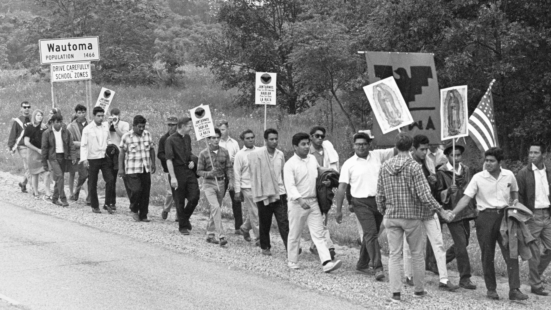 A black-and-white photo of people holding signs and a U.S. flag while marching along a gravel shoulder to a road with a "Wautoma, Population 1466" sign and trees in the background.