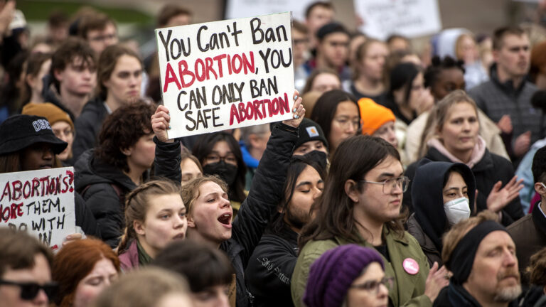 Protestors stand in a crowd with one holding a sign that reads You Can't Ban Abortion, You Can Only Ban Safe Abortion.