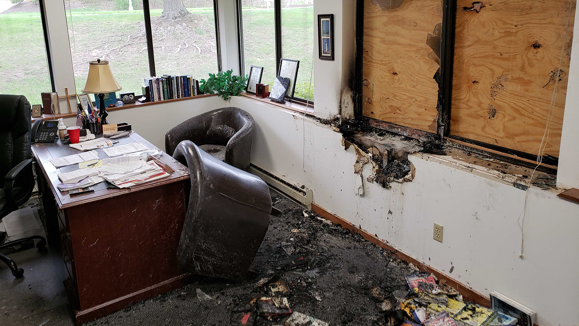 In an office with a desk, multiple chairs and other objects, burned materials and other detritus cover a floor beneath a window frame covered by plywood and surrounded by scorch marks.