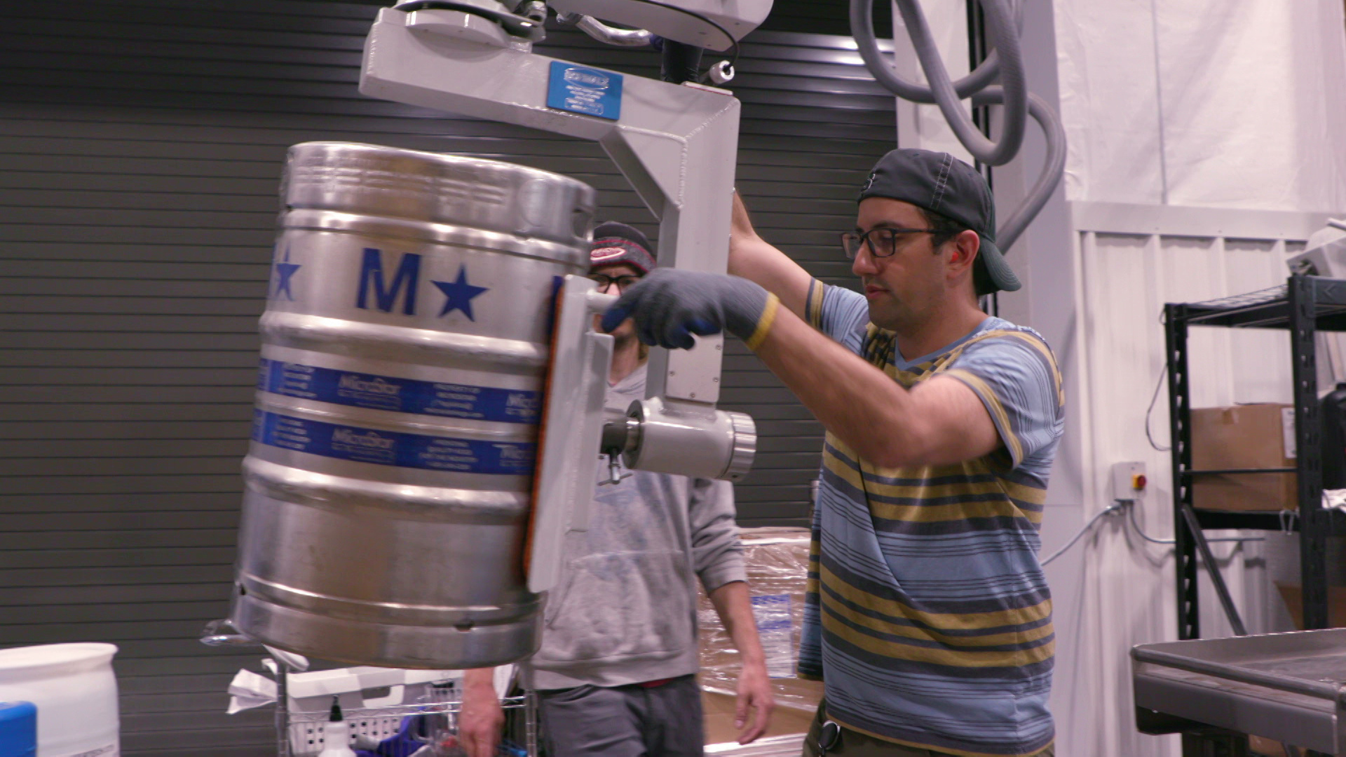 Two brewery workers use a machine mounted overhead to move a keg.