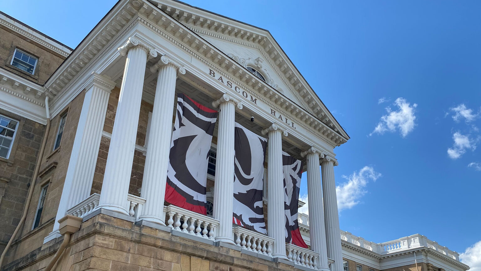 A trio of banners depicting Bucky Badger are placed between pillars on the facade of Bascom Hall on the UW-Madison campus, with a blue sky with a few clouds in the background.