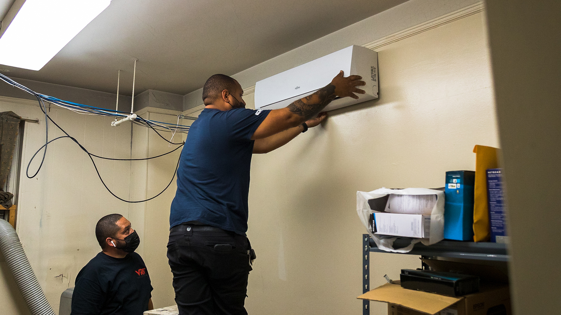 A standing contractor adjusts the placement of a wall-mounted utility appliance inside a building as another contractor watches.