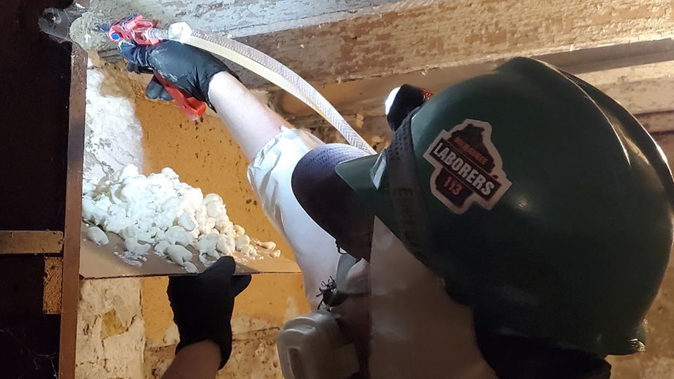 A contractor wearing a safety helmet sprays foam insulation in a basement.