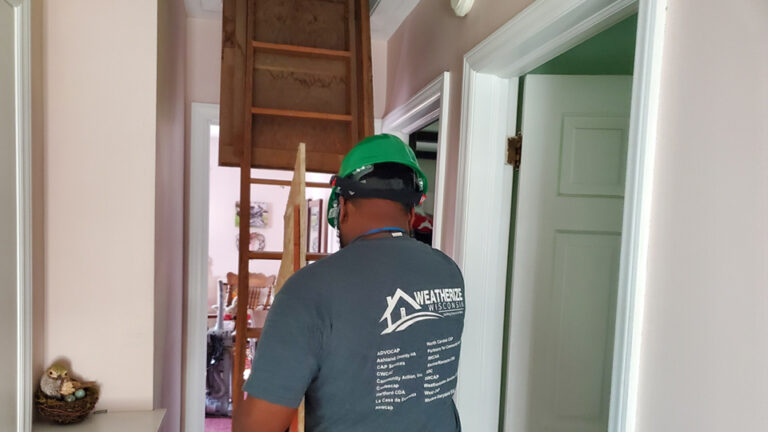 A contractor wearing a safety helmet carries plywood in the hallway of a house while approaching a drop-down ladder extending from an attic entrance.