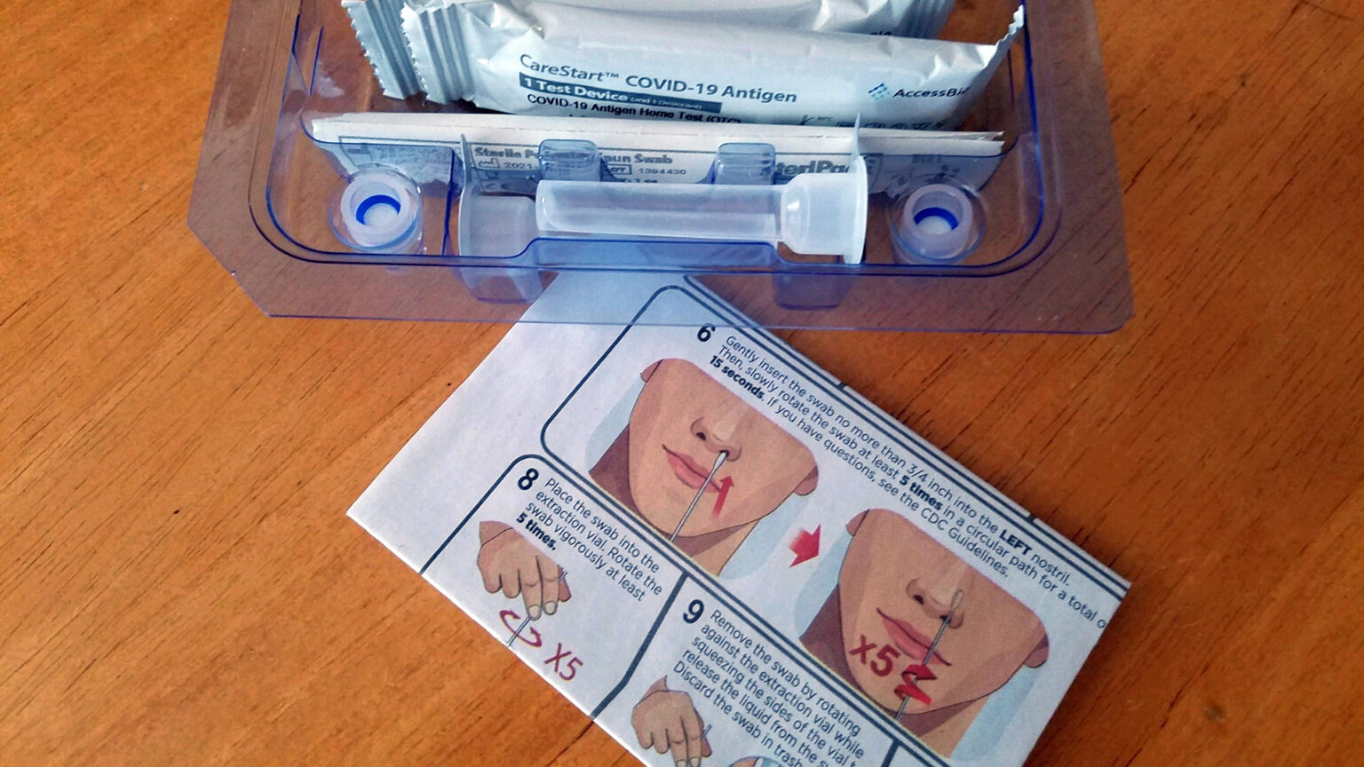 A plastic tray containing COVID-19 testing materials sits next to an instruction manual on top of a wood table.