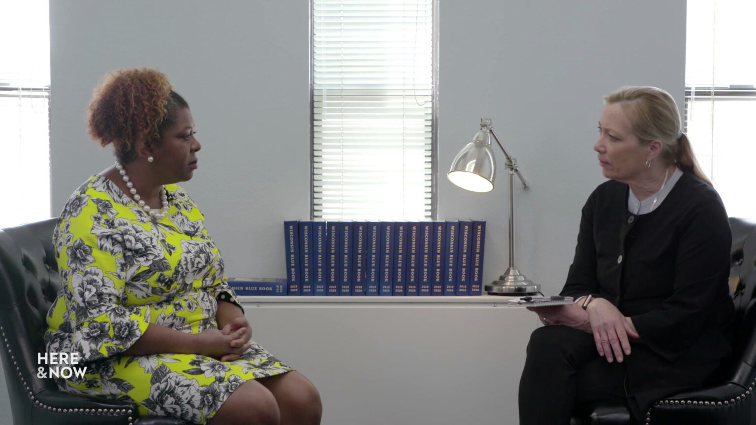 Frederica Freyberg and LaTonya Johnson sit in chairs in a room with Wisconsin Blue Books, a lamp and windows in the background.