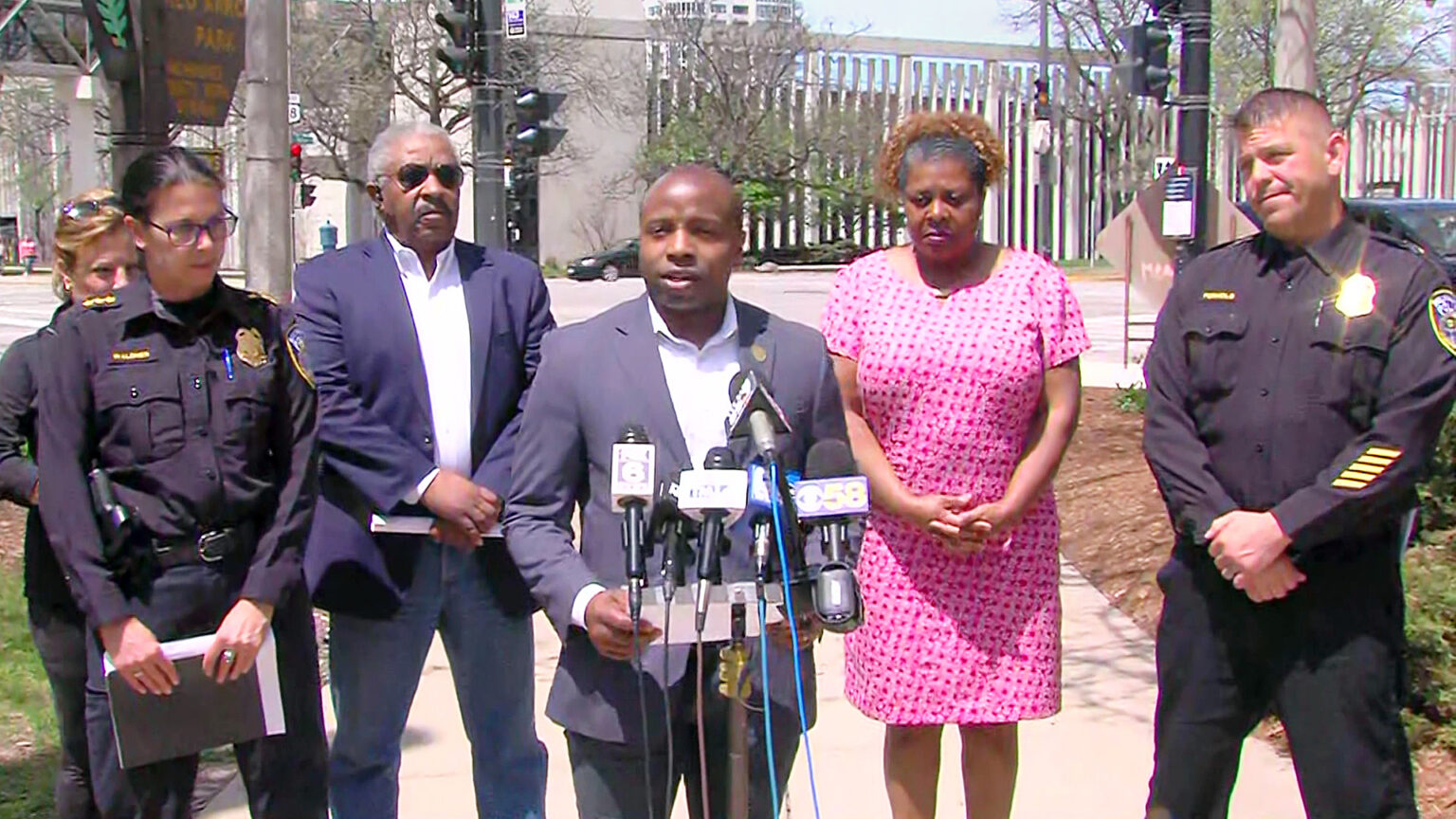 Cavalier Johnson stands behind a podium mounted with multiple news microphones, with police officers and other individuals standing on each side, with an intersection and buildings in the background. 