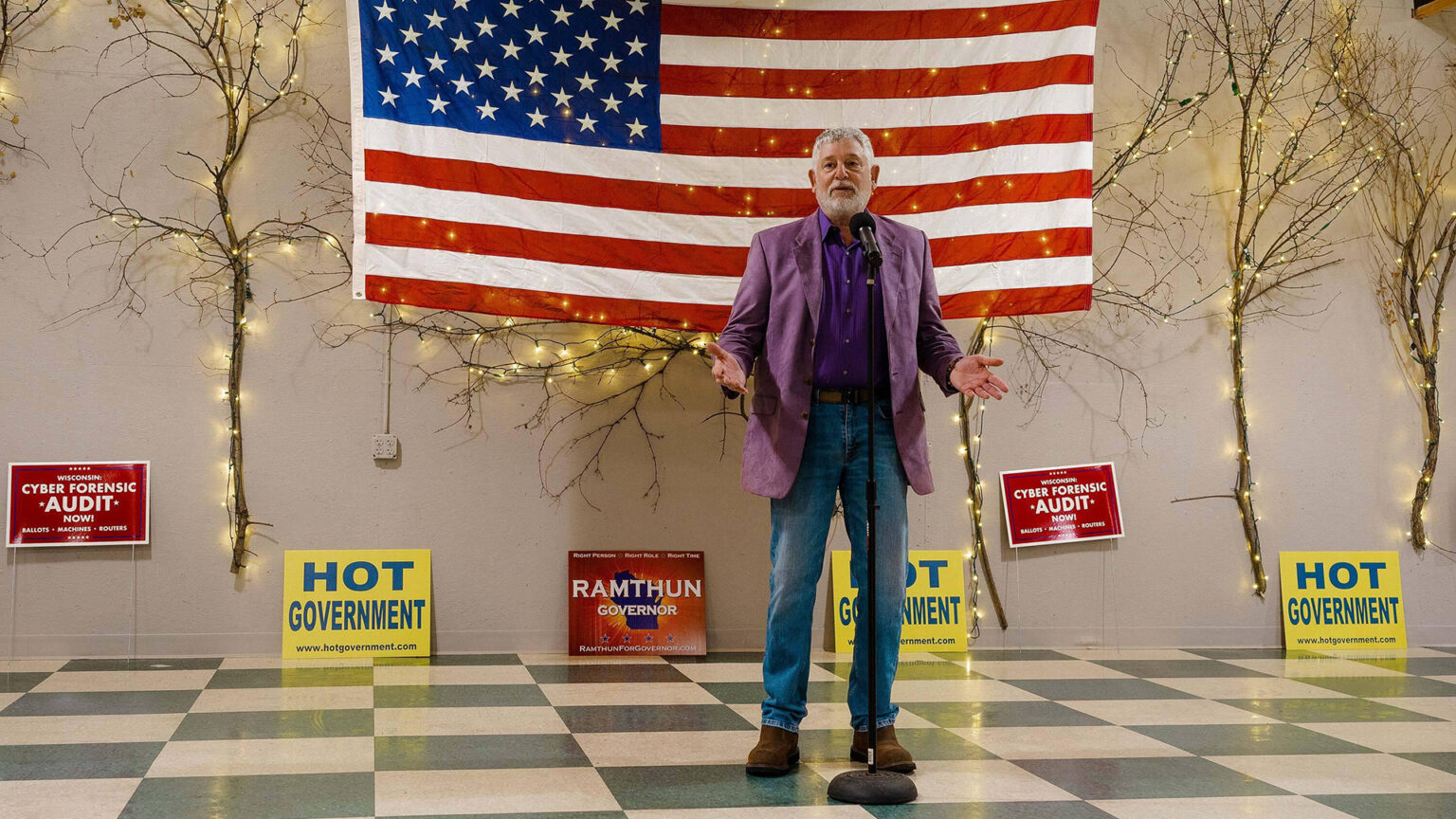 Jay Stone stands in a room speaking into a microphone, with an American flag, political signs and tree branches with LED lights mounted against a wall in the background.