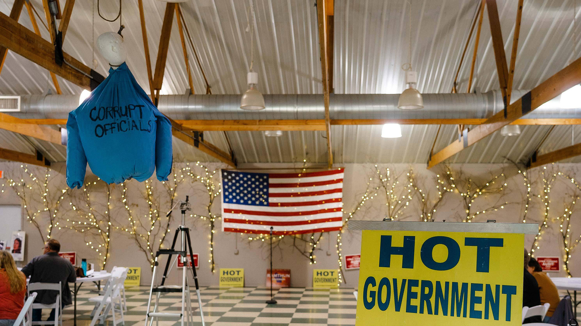 An effigy of a human torso with a shirt labeled "Corrupt Officials" hangs from the wooden rafters of a room with people seated in chairs, an American flag and branches with LED lights mounted on a wall in the background and a political sign reading "HOT Government" in the foreground.