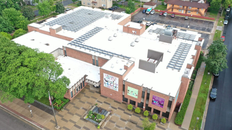 An aerial photo of a brick building with multiple levels and sections shows rows of solar panels on top of its roof, which is white.